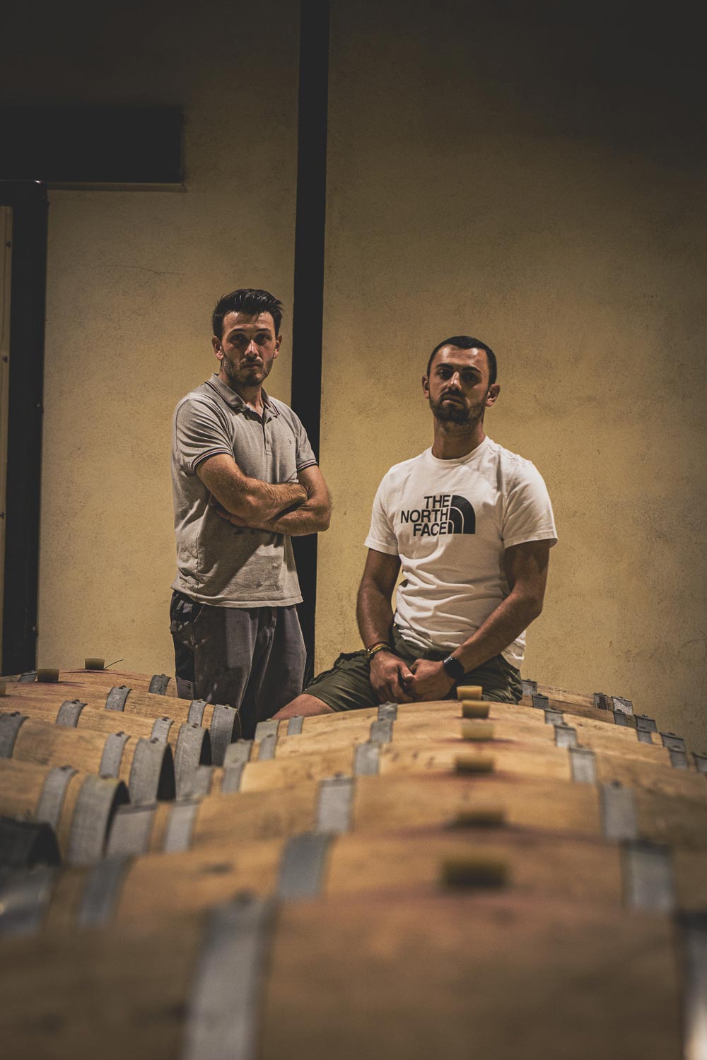 Two men standing and sitting among rows of wooden barrels in a dimly lit cellar.